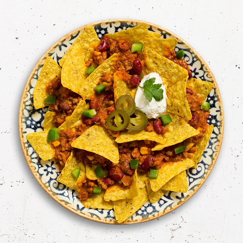 Froyda Chili sin Carne mit Tortilla Chips, grüner Paprika und Jalapenos als Nachos auf blauem Teller hergerichtet.